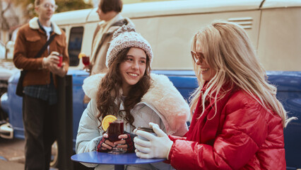 Women chatting with hot drinks at outdoor cafe on cold day. Concept of wellbeing, mindfulness, and seasonal menu advertising evoking warmth and friendly communication.
