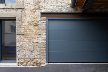 Porte de garage grise anthracite installée par un professionnel, encadrée par un mur en pierre beige et un sol en macadam, entre modernité et authenticité.