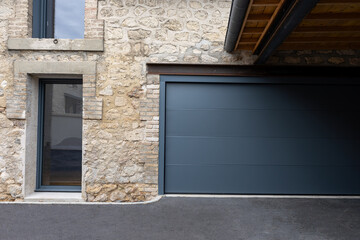 Porte de garage grise anthracite installée par un professionnel, encadrée par un mur en pierre beige et un sol en macadam, entre modernité et authenticité.