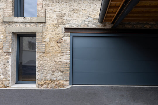 Porte de garage grise anthracite install&eacute;e par un professionnel, encadr&eacute;e par un mur en pierre beige et un sol en macadam, entre modernit&eacute; et authenticit&eacute;.