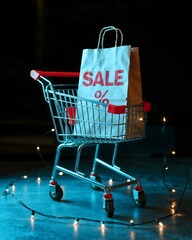 Shopping cart with "SALE %" paper bag and light-bulb garland on dark background