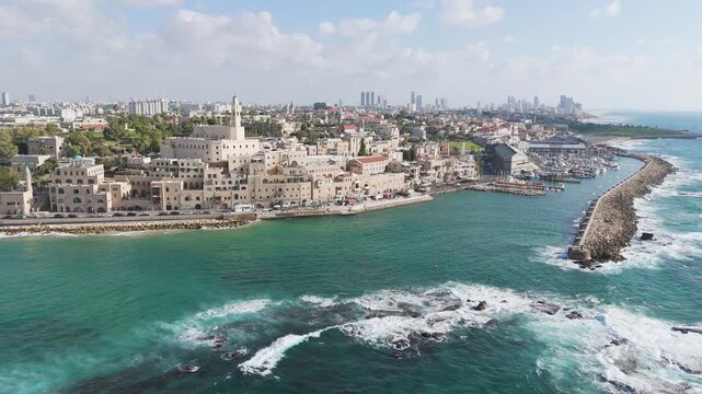 Aerial view of Old Jaffa's sandstone buildings meeting the turquoise Mediterranean Sea, a vibrant contrast against the distant Tel Aviv skyline, Old Jaffa, Tel Aviv-Yafo, Israel.