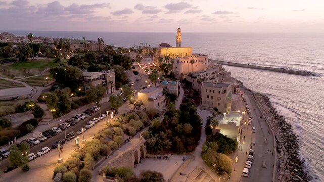 Aerial view of the illuminated St. Peter's Church and old Jaffa buildings juxtaposed against the serene sea at dusk, Old Jaffa, Tel Aviv-Yafo, Israel.