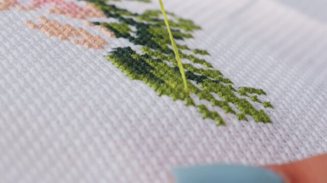 Extreme close up shows crafter passing needle with green thread through cross stitch fabric, shaping leafy pattern on white canvas, crisp weave and texture visible