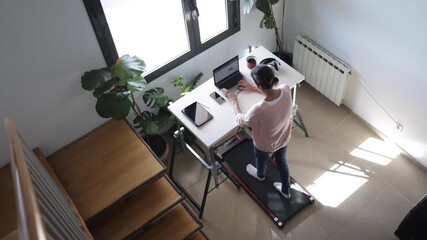 Woman standing and working on a laptop at a treadmill desk in her bright home office, embracing a healthy and active lifestyle while multitasking during work from home