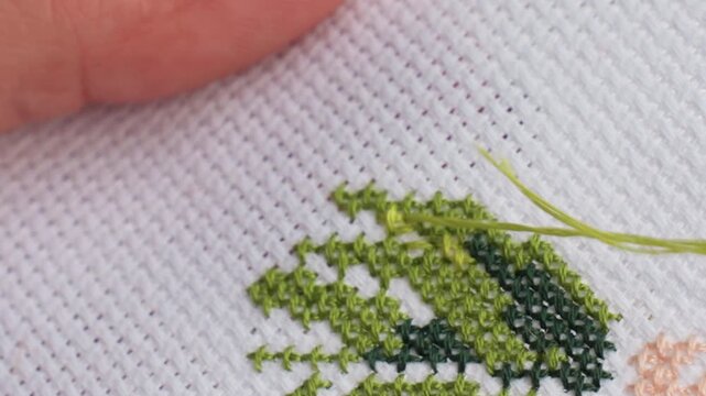 extreme close up skilled crafter passing needle with green thread through cross stitch fabric, precise handwork showing neat x patterns and texture, slow methodical motion