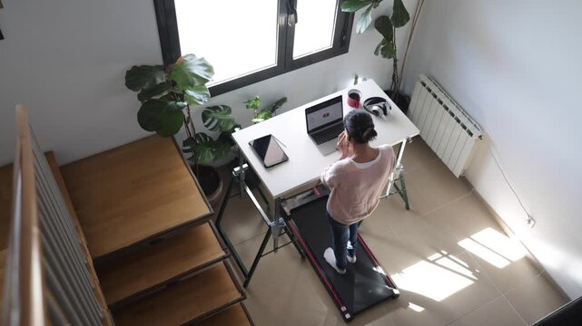 Woman standing and working on a laptop at a treadmill desk in her bright home office, embracing a healthy and active lifestyle while multitasking during work from home