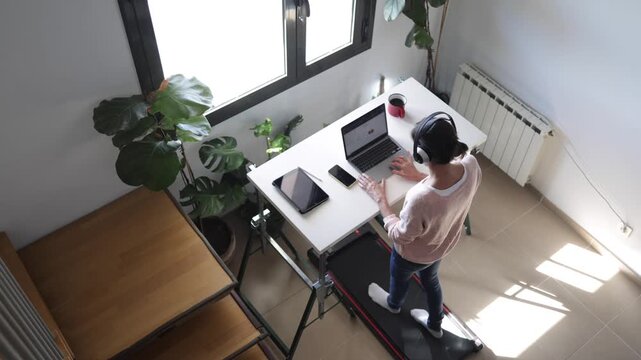 Woman standing and working on a laptop at a treadmill desk in her bright home office, embracing a healthy and active lifestyle while multitasking during work from home