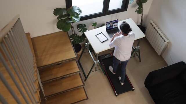 Mother multitasking on a treadmill desk during a video call as her two young sons interrupt and play around her, illustrating the challenge of balancing work and parenting at home