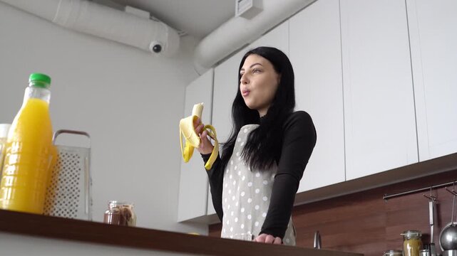 young woman in apron eats banana in the kitchen, girl bites banana and snacks on fruit at home