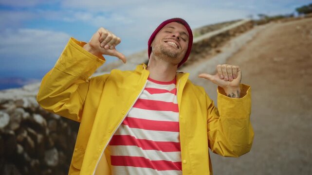 Young man in yellow raincoat and red striped shirt proudly points at himself on a sunny urban street with a mountain landscape background.
