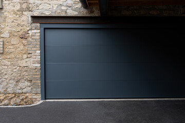 Porte de garage grise anthracite installée par un professionnel, encadrée par un mur en pierre beige et un sol en macadam, entre modernité et authenticité.