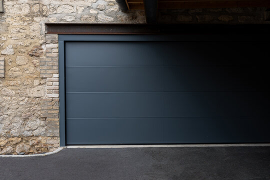 Porte de garage grise anthracite install&eacute;e par un professionnel, encadr&eacute;e par un mur en pierre beige et un sol en macadam, entre modernit&eacute; et authenticit&eacute;.