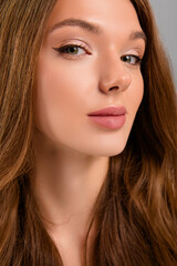 Fototapeta premium Portrait of a young woman with long brown hair showcasing soft makeup and glowing skin on a plain grey background