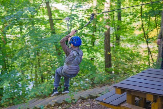 young boy in a blue helmet and harness riding a zip line through a lush green forest adventure park, high-speed outdoor activity descending towards a wooden platform, concept of excitement, speed