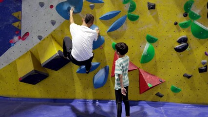 Man spotting boy climbing indoor rock wall at a gym, father and son bonding during a fitness workout session and learning new skills with support and guidance