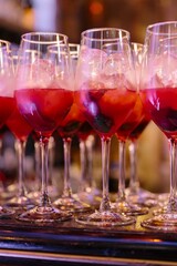 Close-up of a line of bright red Aperol or spritz cocktails with ice and fresh mint leaves in tall, stemmed glasses on a dark reflective surface