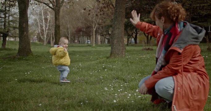 A mother and her young child enjoy a beautiful day at the park. The child takes first steps towards their mom with flowers in hand.