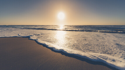 The image shows a beach at sunrise. In the foreground, you can see the wet sand and the foam from a wave that has just broken gently on the shore. The sea occupies much of the scene, with small waves