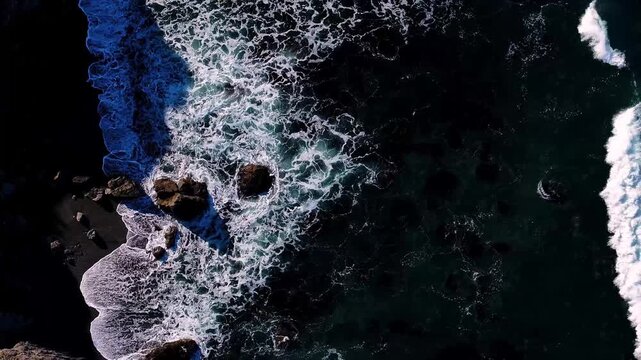 Aerial view of the ocean waves crashing on the dark, rocky California coastline, creating a dynamic interplay of textures and tones, California, United States.