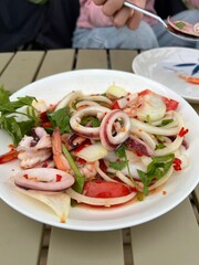 A plate of seafood salad is served on a light slatted table, featuring squid rings, shrimp, onions, tomato wedges, cilantro, and red chili slices. The dish is vibrant and spicy, with a fresh and color