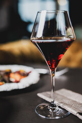A close-up of a glass of red wine on a dark table. The modern, diamond-shaped glass has a slender stem, and a blurred background shows a plate of food, creating an elegant, sophisticated atmosphere.