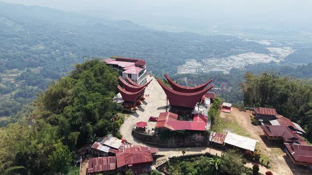 Aerial view of traditional Tongkonan houses with curved roofs on a hillside in Tana Toraja, Indonesia, surrounded by mountains and clouds.	
