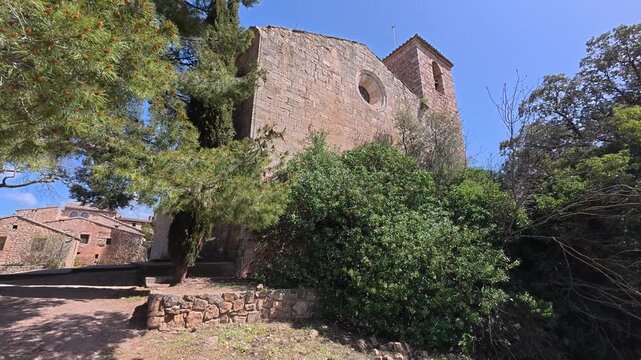 Plano corto en movimiento mostrando la iglesia rom&aacute;nica de Siurana, Tarragona, mientras la c&aacute;mara avanza a pie entre &aacute;rboles y piedra antigua. El video transmite calma, historia y autenticidad.