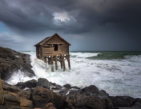 a dilapidated wooden shack stands precariously on rocky shores as massive waves crash around it under a dark stormy sky