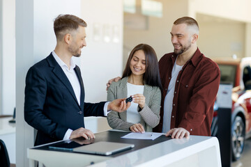 Happy caucasian millennial couple receiving car keys from salesman after signing contract in dealership center, smiling young spouses buying automobile in modern showroom