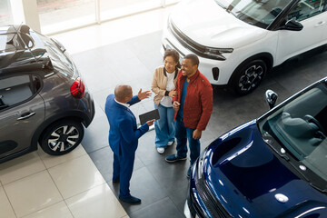 Inside a modern car dealership, a young African American couple engages with a salesman while exploring various vehicles.