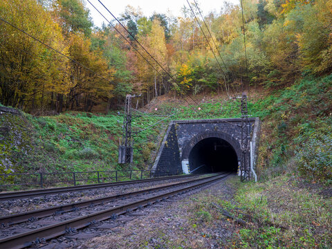Railway tunnel entrance near Margecany, Slovakia, surrounded by autumn forest. Train tracks and overhead wires lead into the stone tunnel in a mountain landscape.