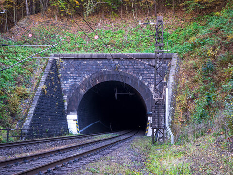 Railway tunnel entrance near Margecany, Slovakia, surrounded by autumn forest. Train tracks and overhead wires lead into the stone tunnel in a mountain landscape.