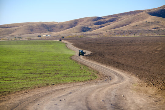 A rural landscape with a dirt road winding between green fields and brown, freshly plowed soil. A tractor moves along the path beneath rolling hills and a clear blue sky