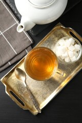 Tasty tea in glass cup, sugar cubes, teaspoon and teapot on black wooden table, flat lay