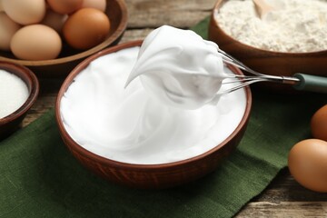 Whipped egg whites in bowl, whisk and ingredients on wooden table, closeup
