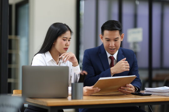 Two business professionals are sitting at a desk, reviewing documents and discussing work during a productive office meeting, utilizing a laptop and paperwork for analysis and collaboration.