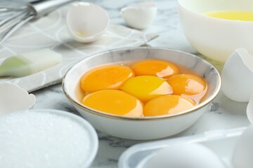 Broken eggs, yolks, whites, sugar, brush and whisk on white marble table, closeup