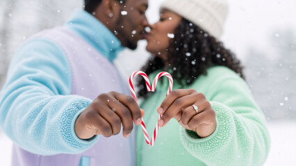 Close-up shot focuses on the hands of a Black couple holding two red and white striped candy canes to form heart. A romantic black couple kissing in the snow while holding candy canes in heart shape.