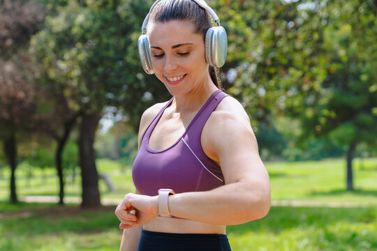 Active woman wearing wireless headphones and sportswear, smiling while checking smartwatch after training in green park. Outdoor fitness, healthy lifestyle and modern technology.