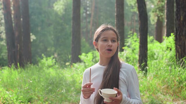 Teenage girl holding tea cup and eating breadsticks while standing in green forest, sunlight softly illuminating her thoughtful face as she looks around lost in quiet reflection
