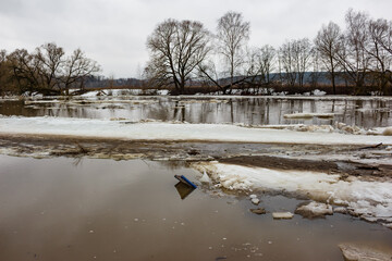 A blue, leaning sign partially submerged in a muddy river, pushed by ice floes during a spring ice drift. Bare trees line the far bank under a dull sky