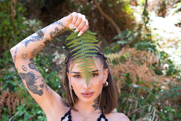 Portrait of a beautiful young woman covering half her face with a green fern leaf. The woman is...