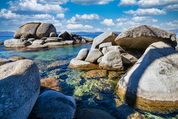 Rocky shoreline at Sand Harbor State Park Lake Tahoe Nevada USA with clear turquoise water sparkling in sunlight and granite boulders along pine forest coast under blue sky with white clouds