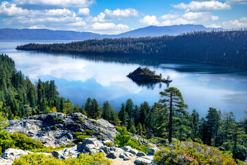 Scenic view of Emerald Bay State Park at Lake Tahoe California USA with Fannette Island pine forest and Sierra Nevada mountains reflecting in calm blue water under bright sky with fluffy white clouds