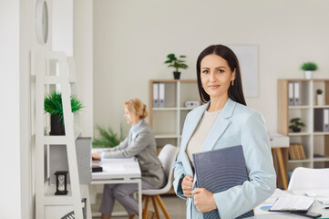 Successful woman in elegant clothing, holding notepad and posing in office room, finished work in...