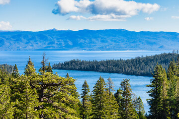 Bright daytime view of Emerald Bay and Lake Tahoe California USA with clear blue water surrounded by green pine forest and Sierra Nevada mountains under sunny sky with scattered white clouds