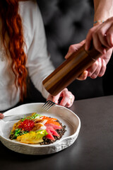 A chef using a pepper mill to season a gourmet dish of salmon and salad at a fine dining restaurant, with a person seated at the table ready to eat