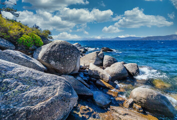 Lake Tahoe California USA scenic rocky shoreline with clear blue water and large granite boulders under bright sky and white clouds summer view of pristine Sierra Nevada nature and landscape