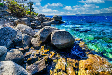 Lake Tahoe California USA scenic rocky shoreline with clear blue water and large granite boulders under bright sky and white clouds summer view of pristine Sierra Nevada nature and landscape